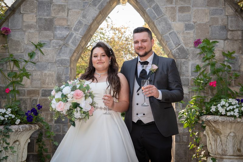 A bride and groom smile under a stone archway, holding champagne flutes surrounded by greenery and flowers.