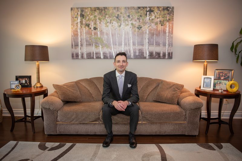 A man in a suit smiles at the camera in a living room decorated with symmetrical side tables and a birch tree painting.