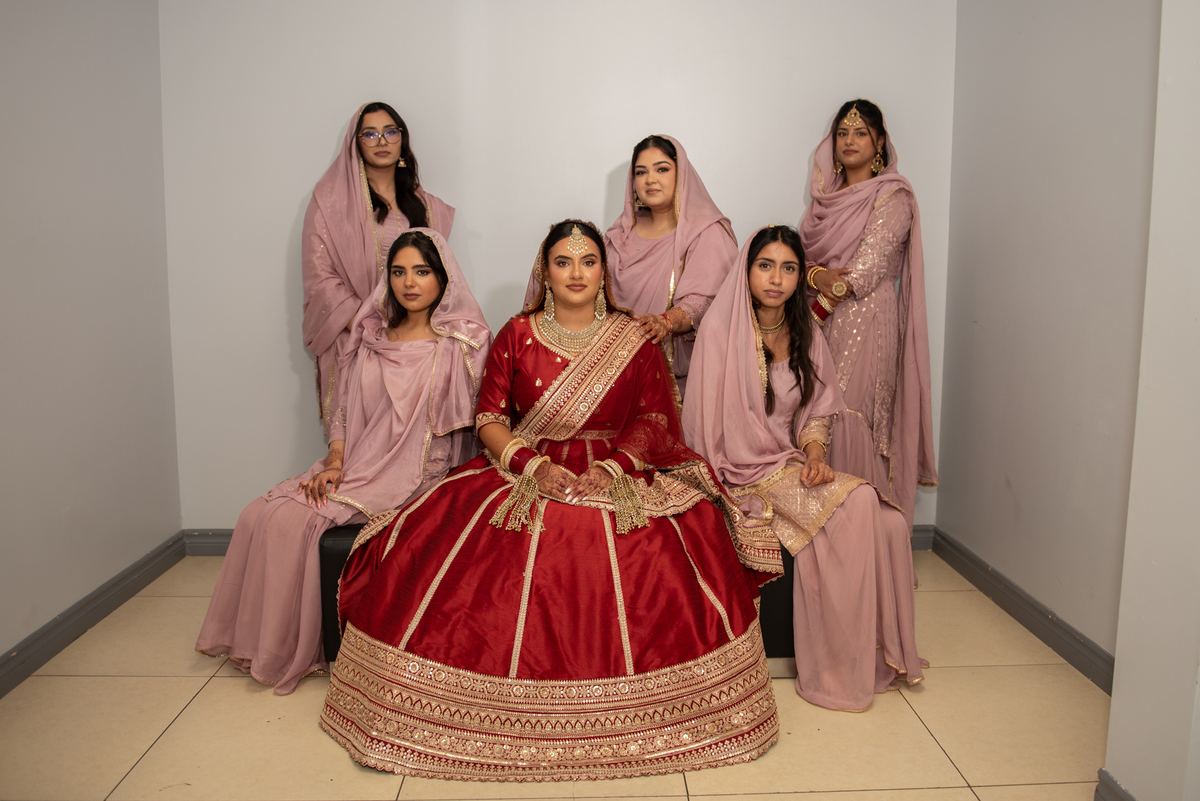 A bride in a red and gold lehenga sits with her pastel-clad bridesmaids for a formal portrait in an indoor setting.