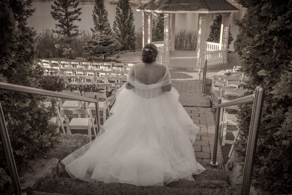 A bride in a voluminous white gown descends stone steps toward an outdoor ceremony framed by trees and chairs.