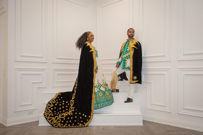 A couple in ornate black velvet and gold embroidery stand smiling on a white staircase during a formal event.