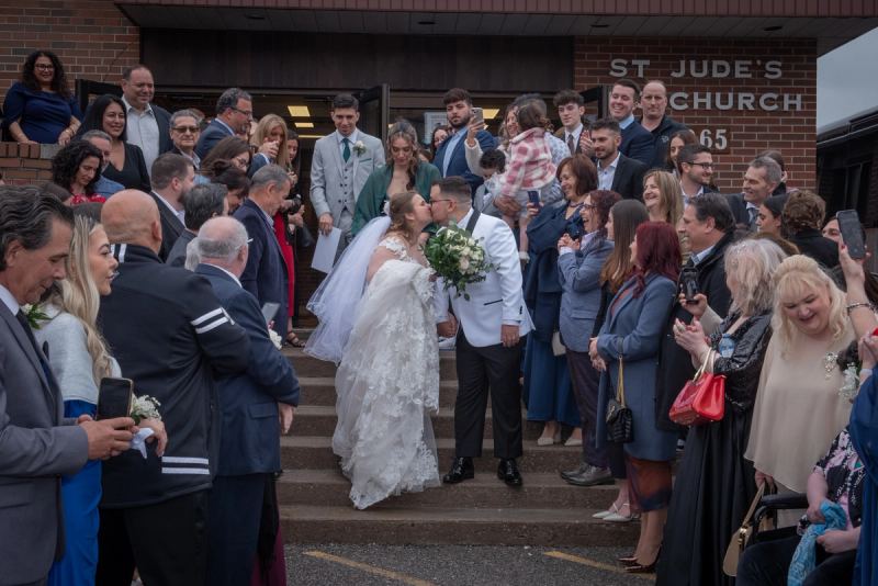 A bride and groom kiss as they descend church steps, surrounded by a smiling crowd holding phones and cameras.