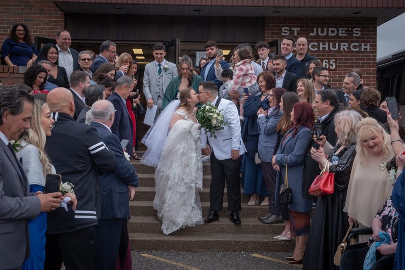 A bride and groom kiss as they descend church steps, surrounded by a smiling crowd holding phones and cameras.