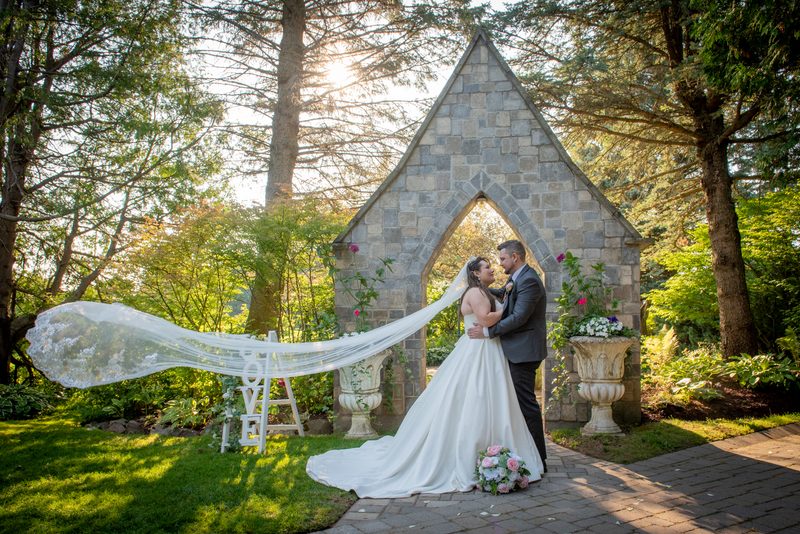 A bride and groom embrace beneath a stone Gothic archway in a sun-dappled garden with floral arrangements nearby.