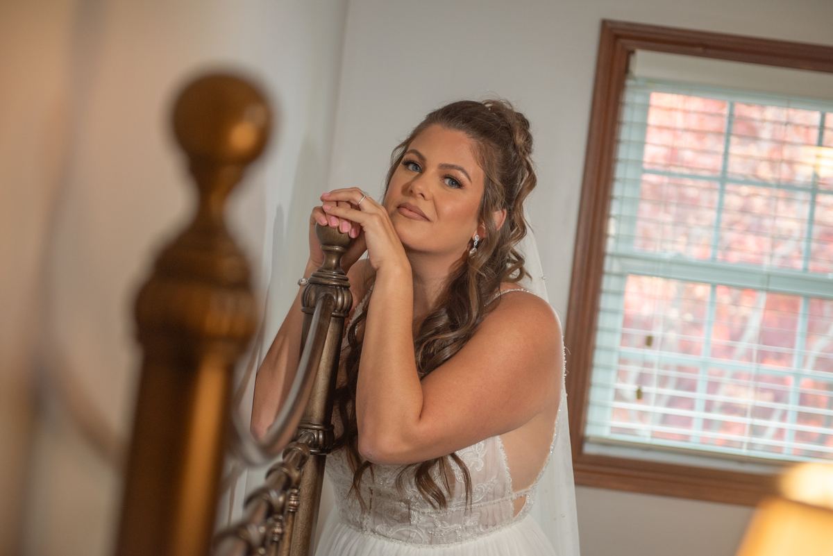 A bride in a white gown stands by an ornate staircase, hands resting on the railing, gazing thoughtfully upward.