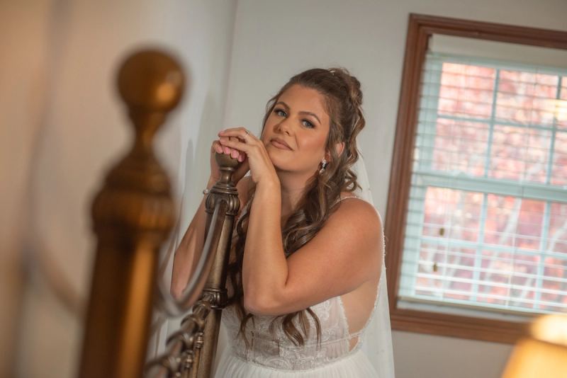 A bride in a white gown stands by an ornate staircase, hands resting on the railing, gazing thoughtfully upward.