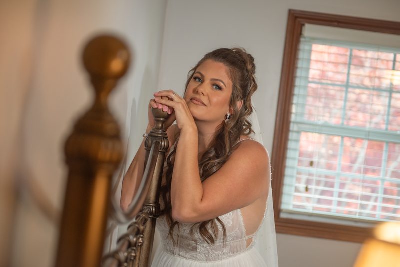 A bride in a white gown stands by an ornate staircase, hands resting on the railing, gazing thoughtfully upward.