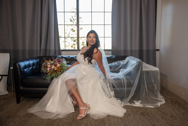 A bride sits gracefully on a black leather couch, smiling warmly at the camera while holding a colorful bouquet.