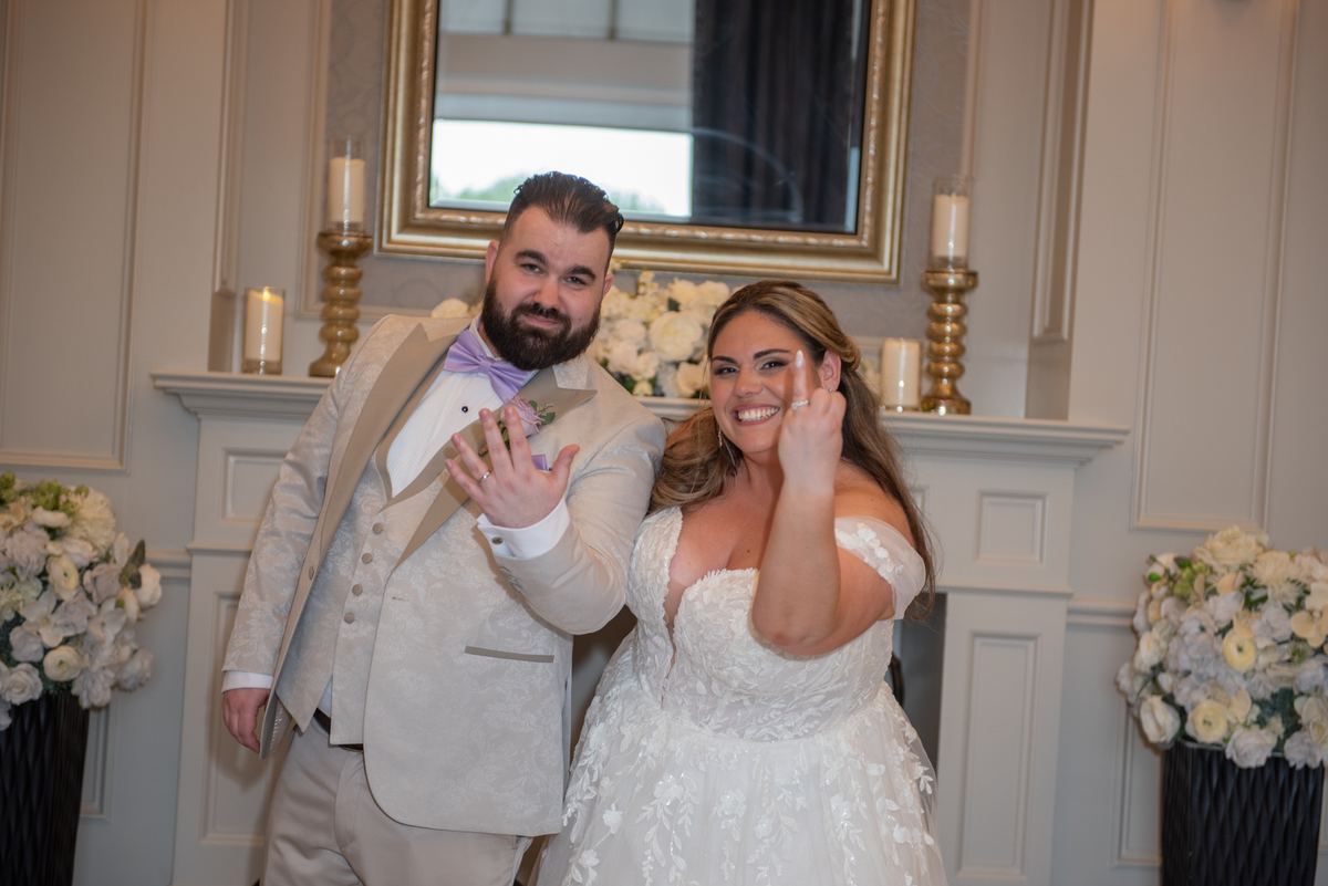 A smiling bride and groom show their wedding rings in front of a fireplace with candles and flowers.