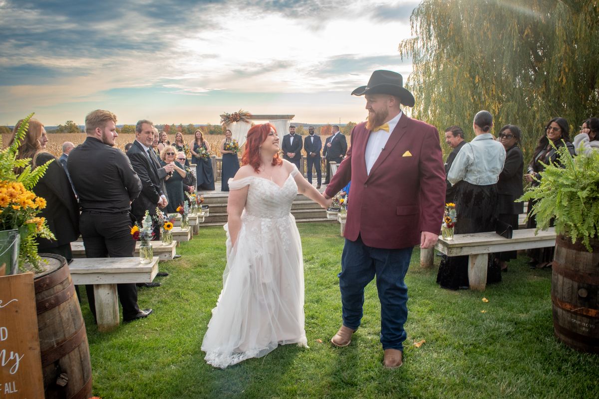 A bride in white and groom in a cowboy hat walk hand-in-hand down an outdoor aisle lined with guests.