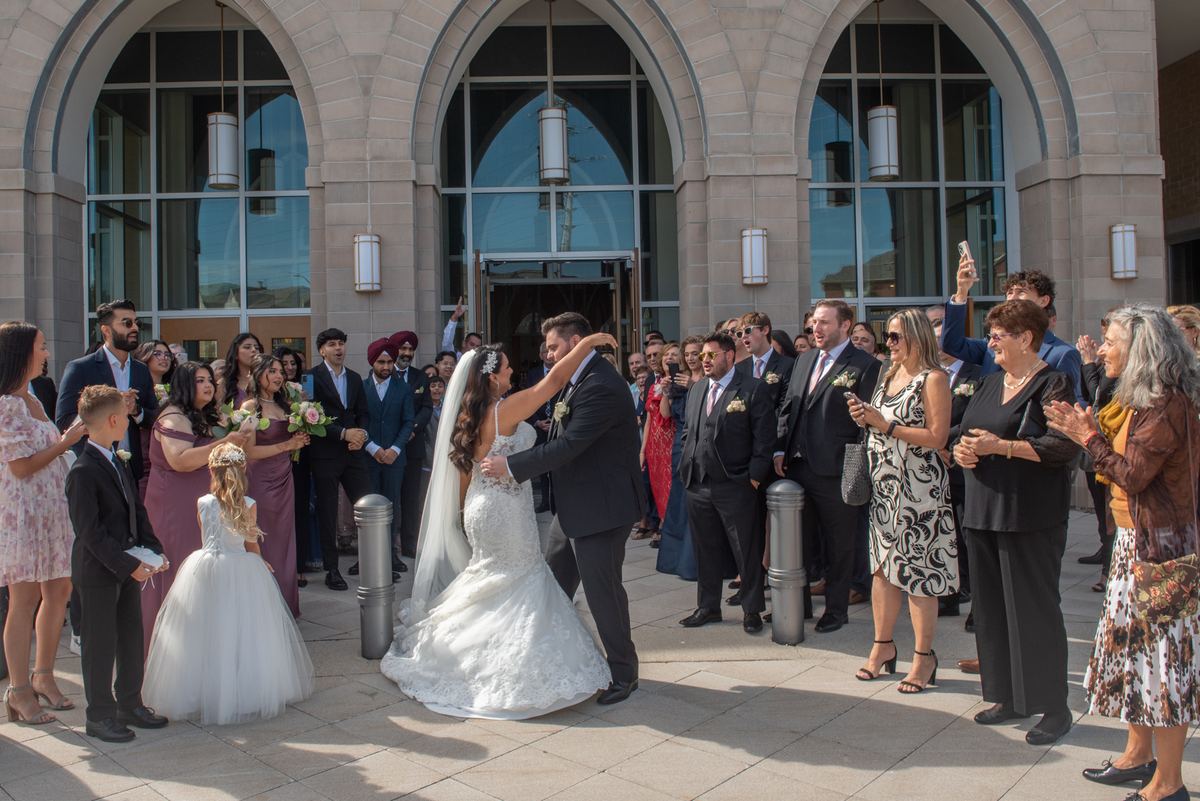 A bride and groom dance outside a modern stone building as guests clap, illuminated by warm sunlight.