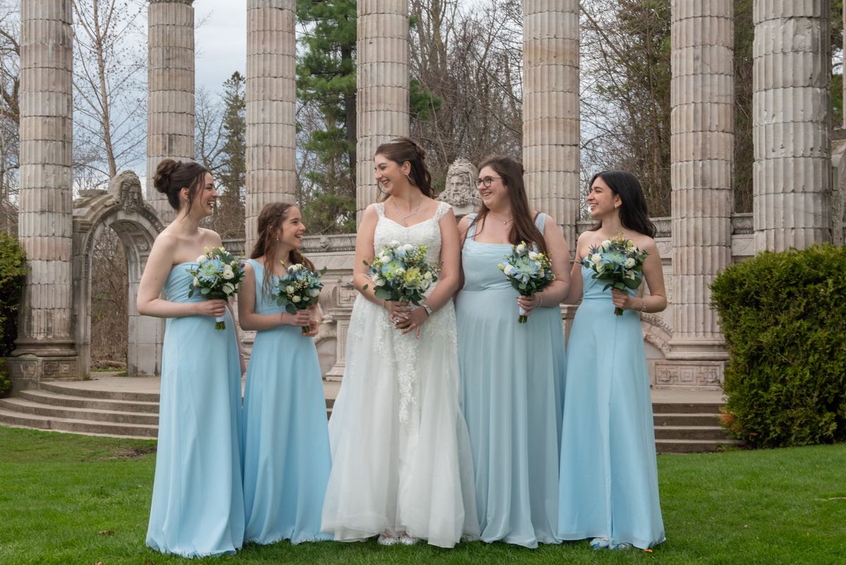 A bride and four bridesmaids in pastel dresses stand joyfully under stone columns with bouquets of blue and white flowers.