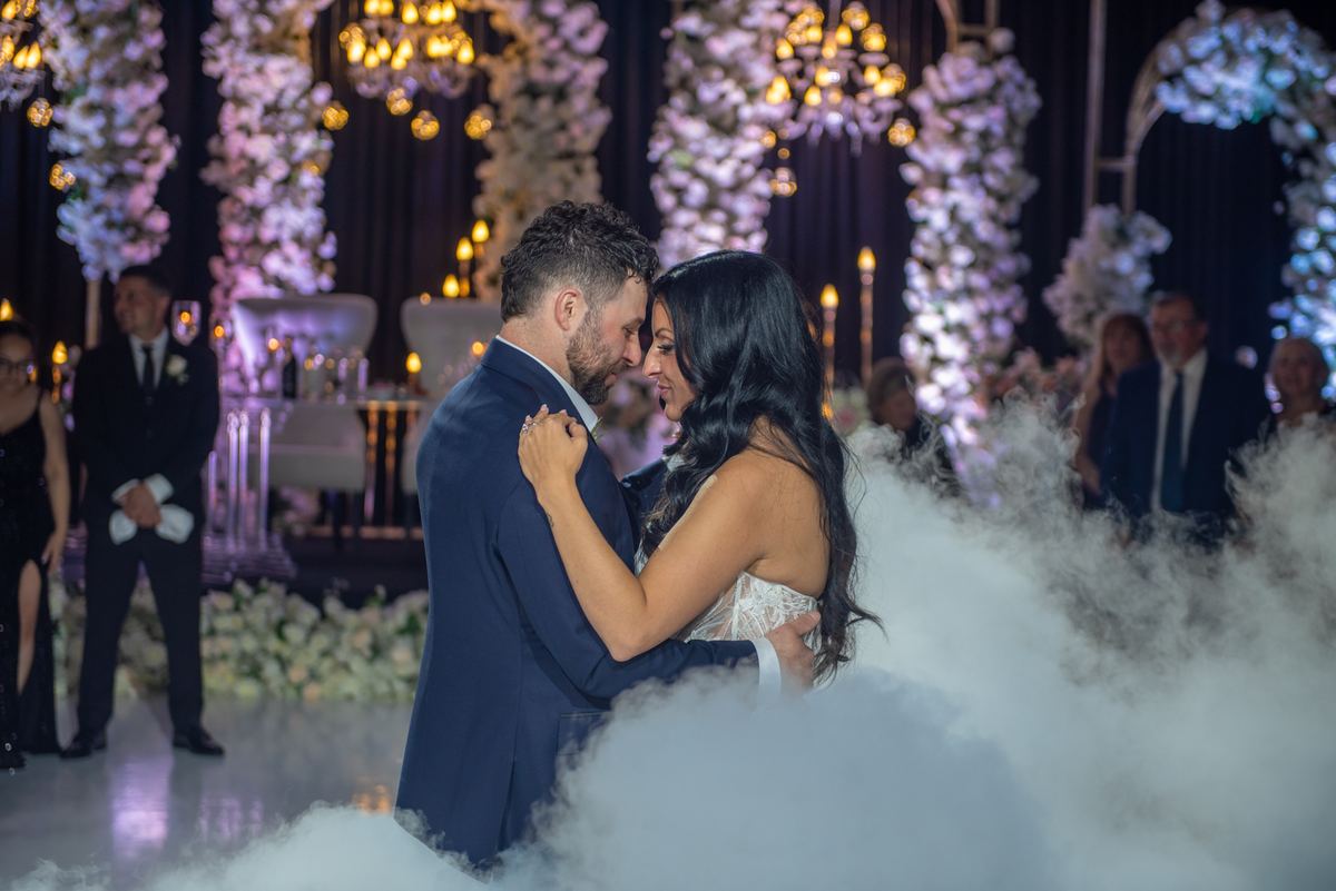Bride and groom dance intimately in foggy, dreamlike setting with blurred guests and glowing chandeliers.