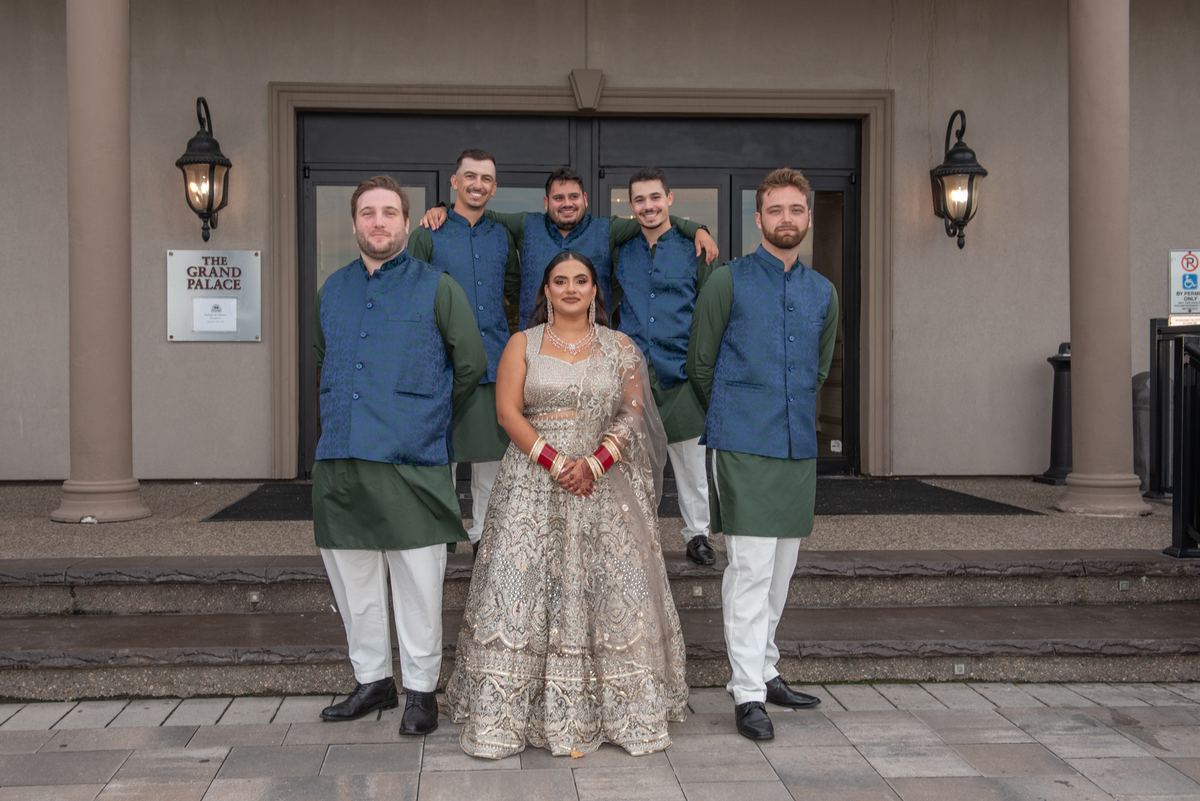 A bride in a silver lehenga and six groomsmen in matching sherwanis pose on palace steps, illuminated by warm lanterns.