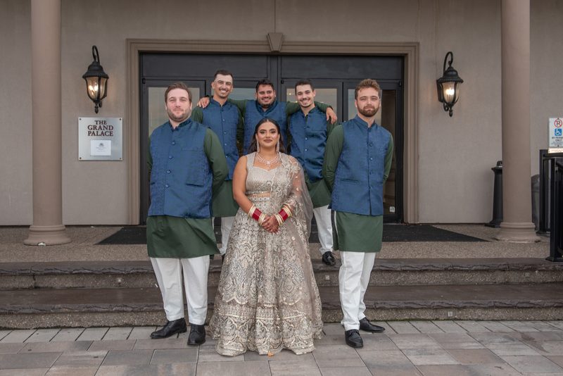 A bride in a silver lehenga and six groomsmen in matching sherwanis pose on palace steps, illuminated by warm lanterns.
