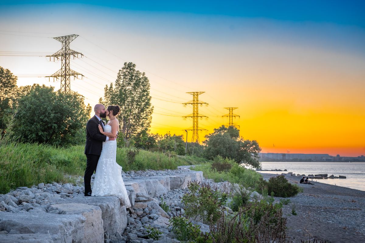 A newlywed couple kisses passionately on a rocky shore at sunset, silhouetted against an orange and blue sky.