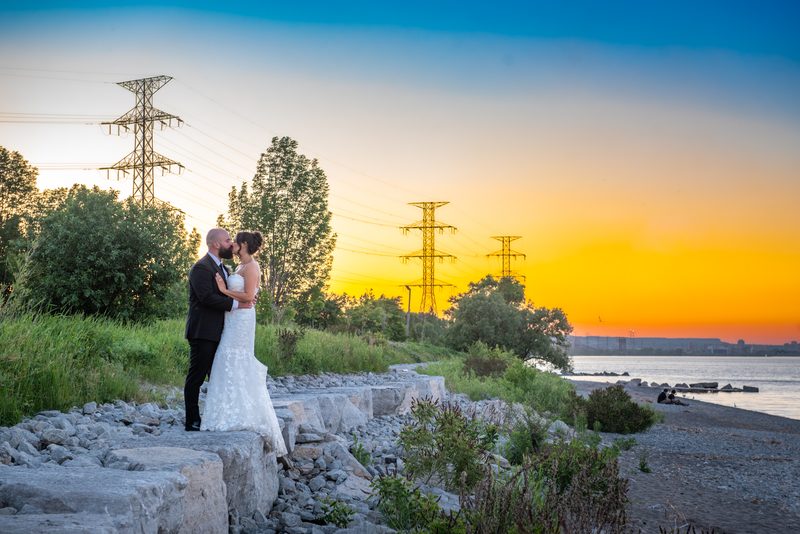 A newlywed couple kisses passionately on a rocky shore at sunset, silhouetted against an orange and blue sky.