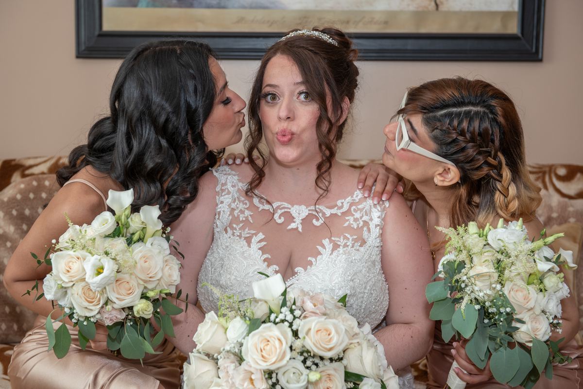 A bride in a lace gown laughs as bridesmaids whisper secrets and hold rose bouquets, creating an intimate moment.