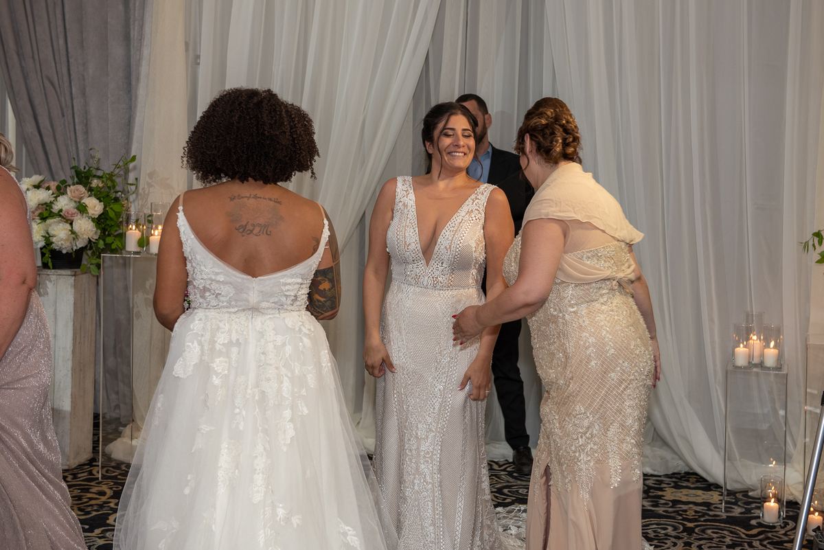 Two brides exchange glances and a touch from a third woman in a candlelit, draped room with floral arrangements.