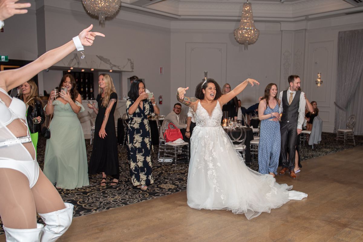 The bride dances joyfully in a grand ballroom, surrounded by cheering guests under warm chandeliers.