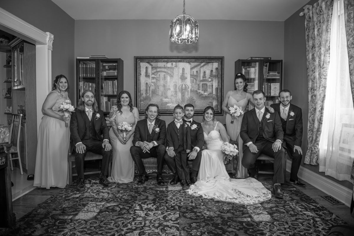 A formal indoor wedding party poses, with the bride and groom seated center amid vintage furnishings and soft light.