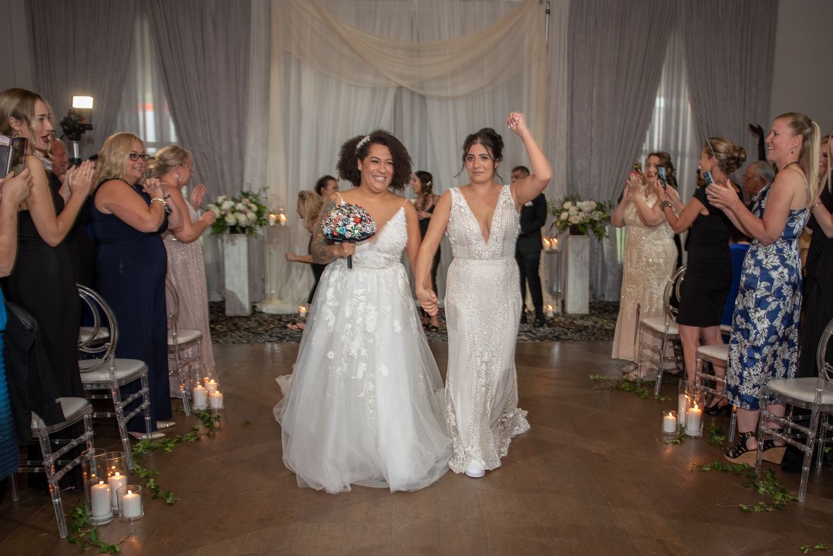 Two brides walk down the aisle hand-in-hand, smiling as guests clap and cheer, one holding a bouquet.