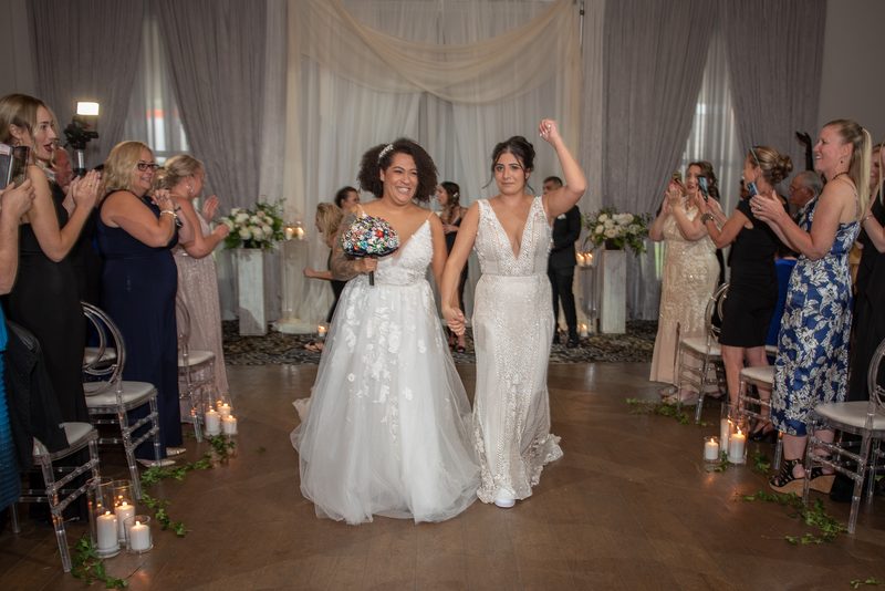 Two brides walk down the aisle hand-in-hand, smiling as guests clap and cheer, one holding a bouquet.