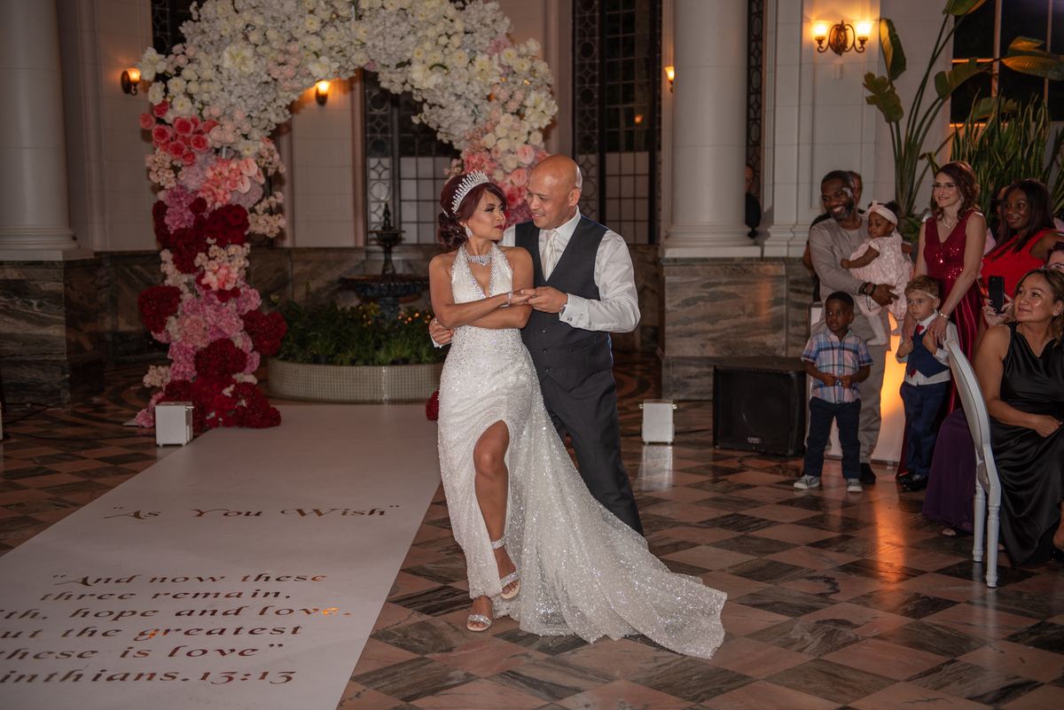 A bride and groom dance in a grand ballroom, surrounded by guests and adorned with floral decorations and scripture banners.