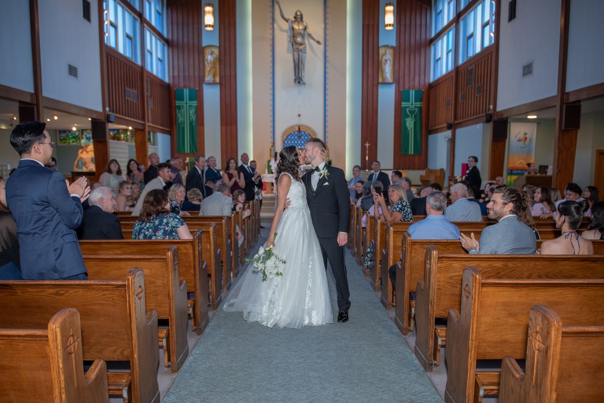 A newlywed couple kisses as guests applaud in a church with wooden pews and a statue of Jesus at the altar.