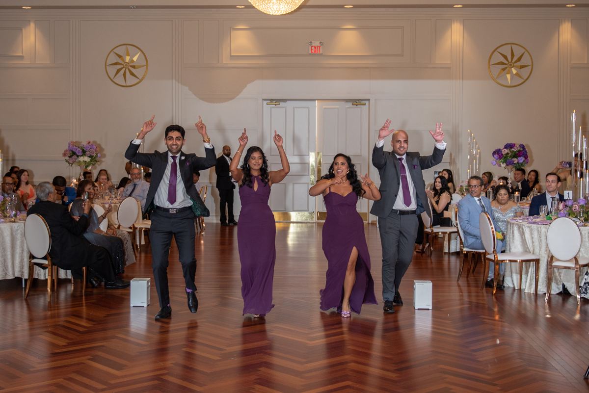 Four guests dance joyfully in a ballroom during a celebration, arms raised, as others watch and cheer.
