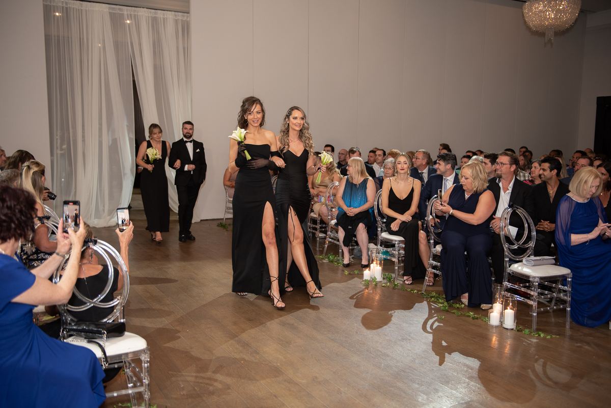 Two women in black gowns walk down a candlelit aisle toward an audience, each holding white flowers.