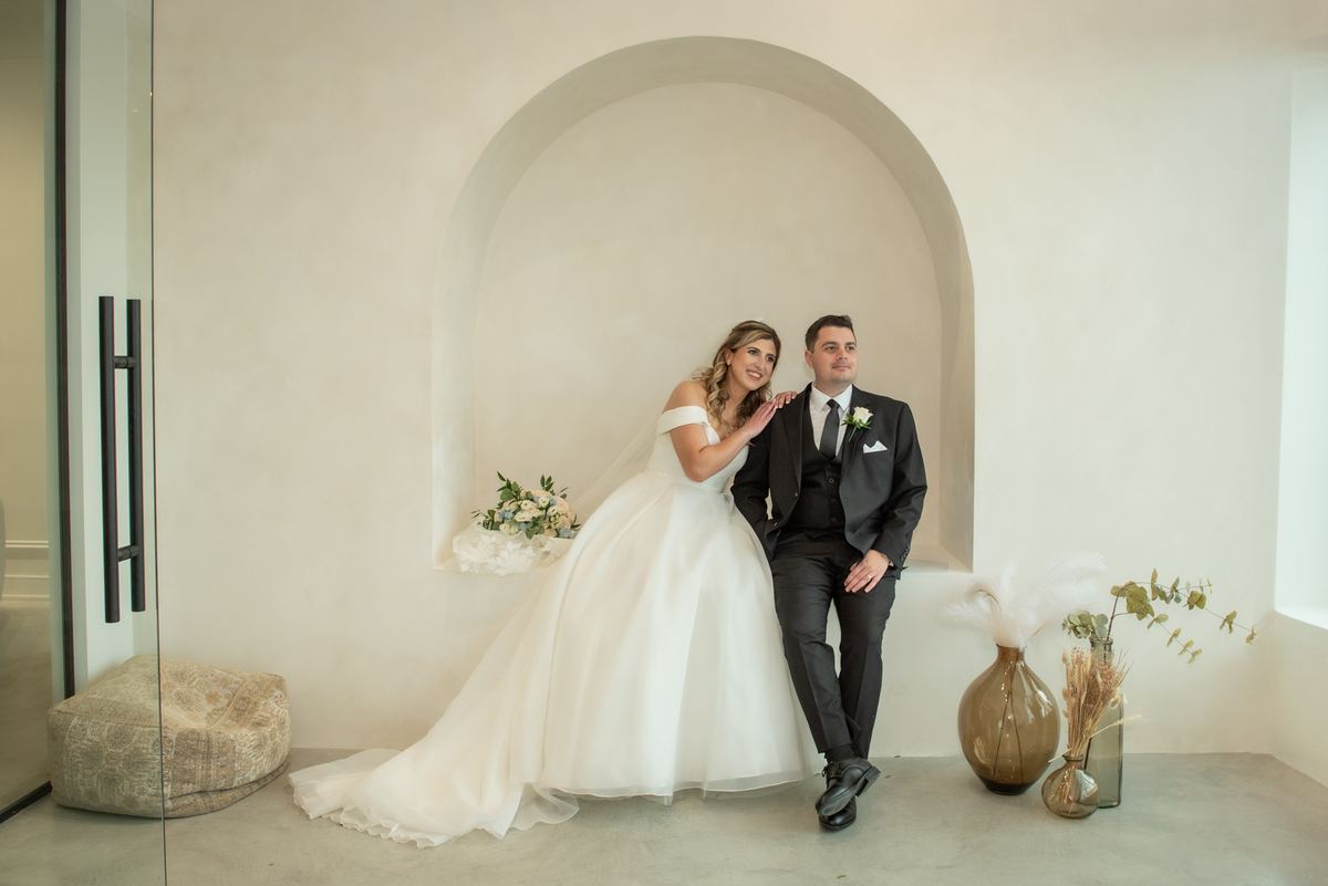 Bride and groom smile warmly in an arched alcove with modern decor and dried flowers, illuminated by soft natural light.