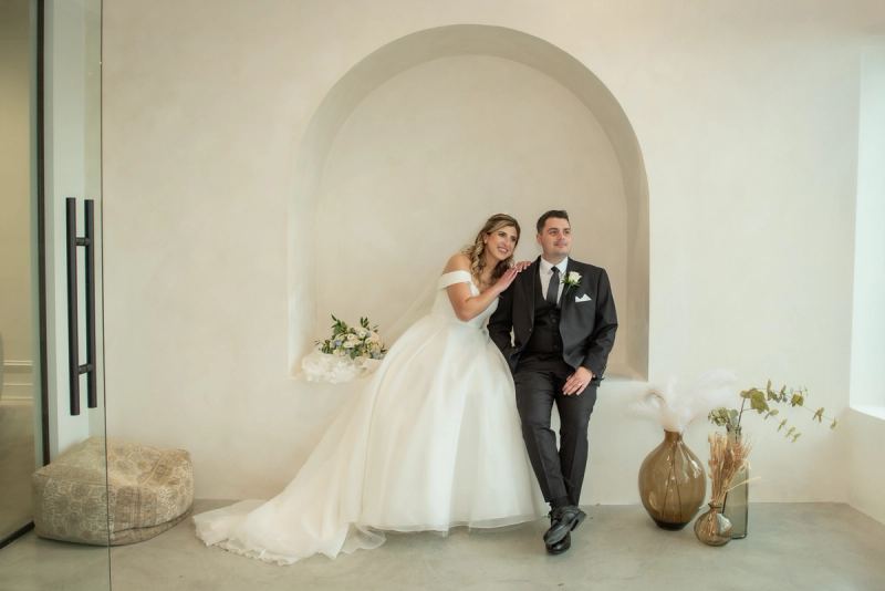 Bride and groom smile warmly in an arched alcove with modern decor and dried flowers, illuminated by soft natural light.