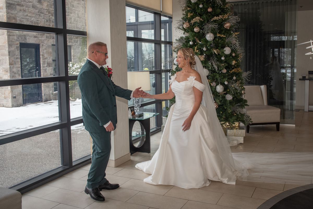 A bride and groom in formal attire share a joyful handshake near a window with snow, under a decorated Christmas tree.