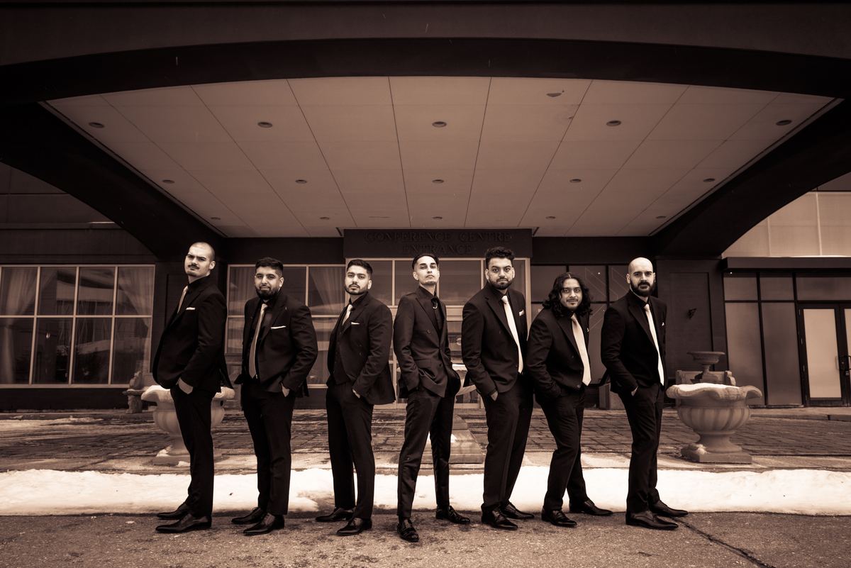 Seven formally dressed men stand outside a modern building in snow, conveying readiness and solemnity.