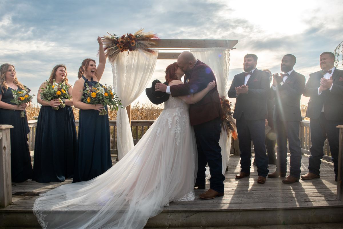 Bride and groom kiss under wooden arch as wedding party cheers, sunlight creates lens flare on deck.