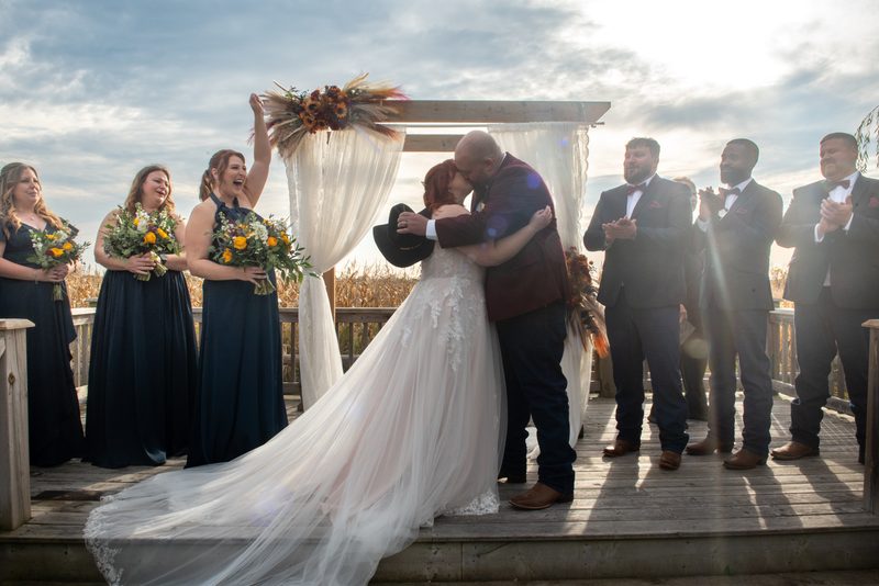 Bride and groom kiss under wooden arch as wedding party cheers, sunlight creates lens flare on deck.