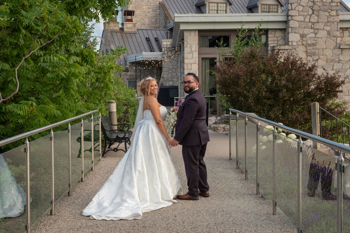 A smiling newlywed couple stands hand-in-hand on a gravel walkway surrounded by greenery and string lights.