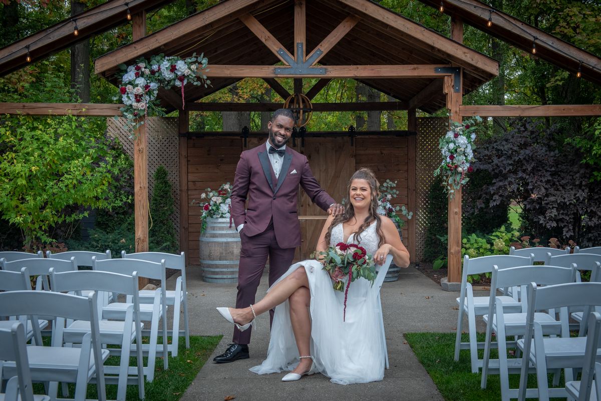 Bride and groom pose joyfully under a wooden arbor adorned with flowers, surrounded by white chairs.