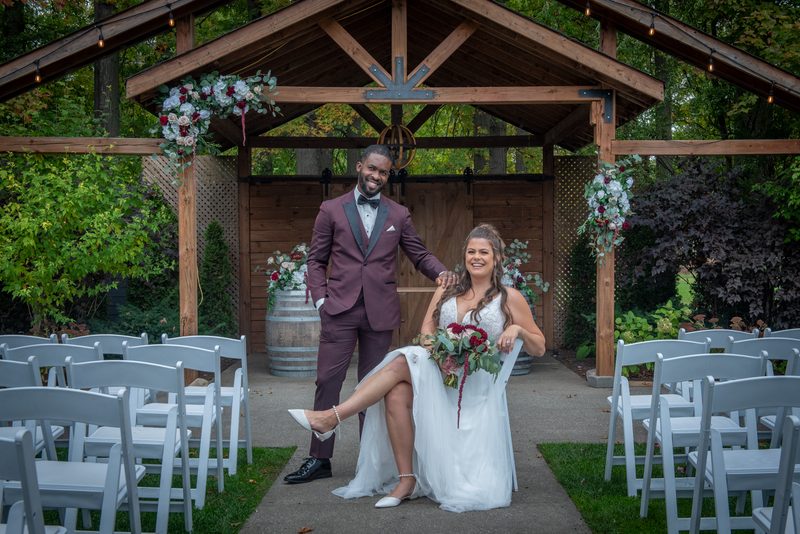 Bride and groom pose joyfully under a wooden arbor adorned with flowers, surrounded by white chairs.