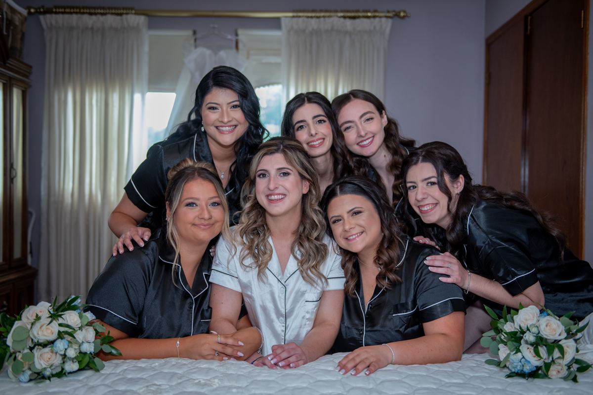 A bride and her bridesmaids smile warmly on a bed, surrounded by soft natural light from sheer curtains.