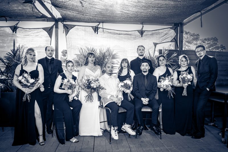 A black-and-white wedding party photo under a canopy with the bride and groom at center surrounded by bridesmaids.