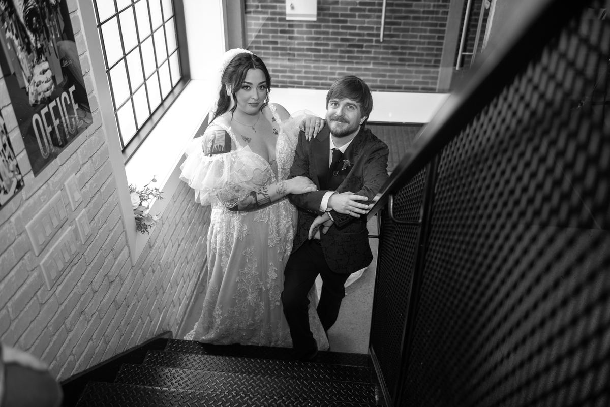 Bride in lace gown and groom descend metal staircase in industrial building, illuminated by natural light.