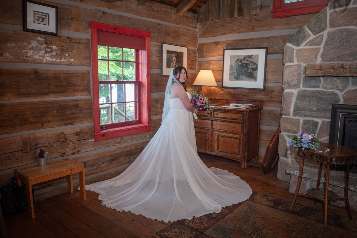 A bride in a white gown stands in a rustic cabin, smiling and holding a bouquet near a red-framed window.