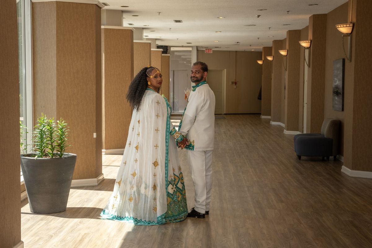 Bride and groom stand in a sunlit hotel corridor, smiling at the camera in front of a large window with wooden floors.