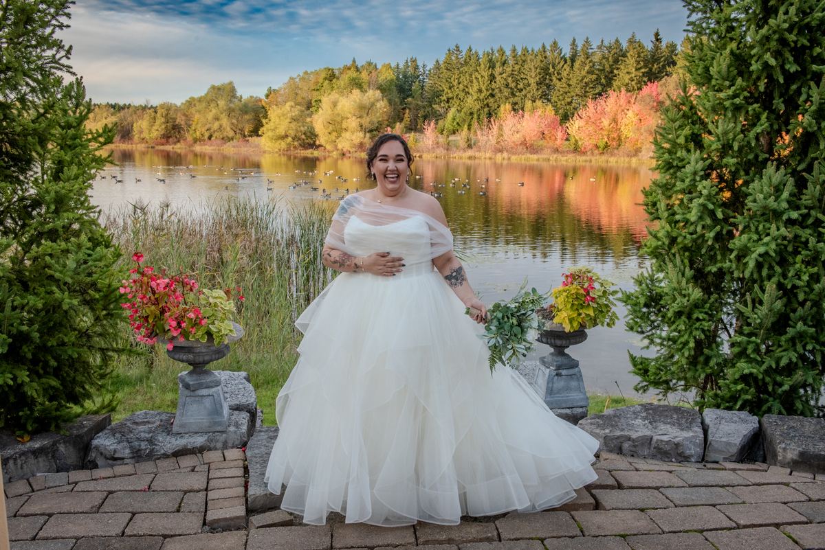 A smiling bride in a white gown stands on a stone patio by a tranquil lake surrounded by autumn foliage and potted flowers.