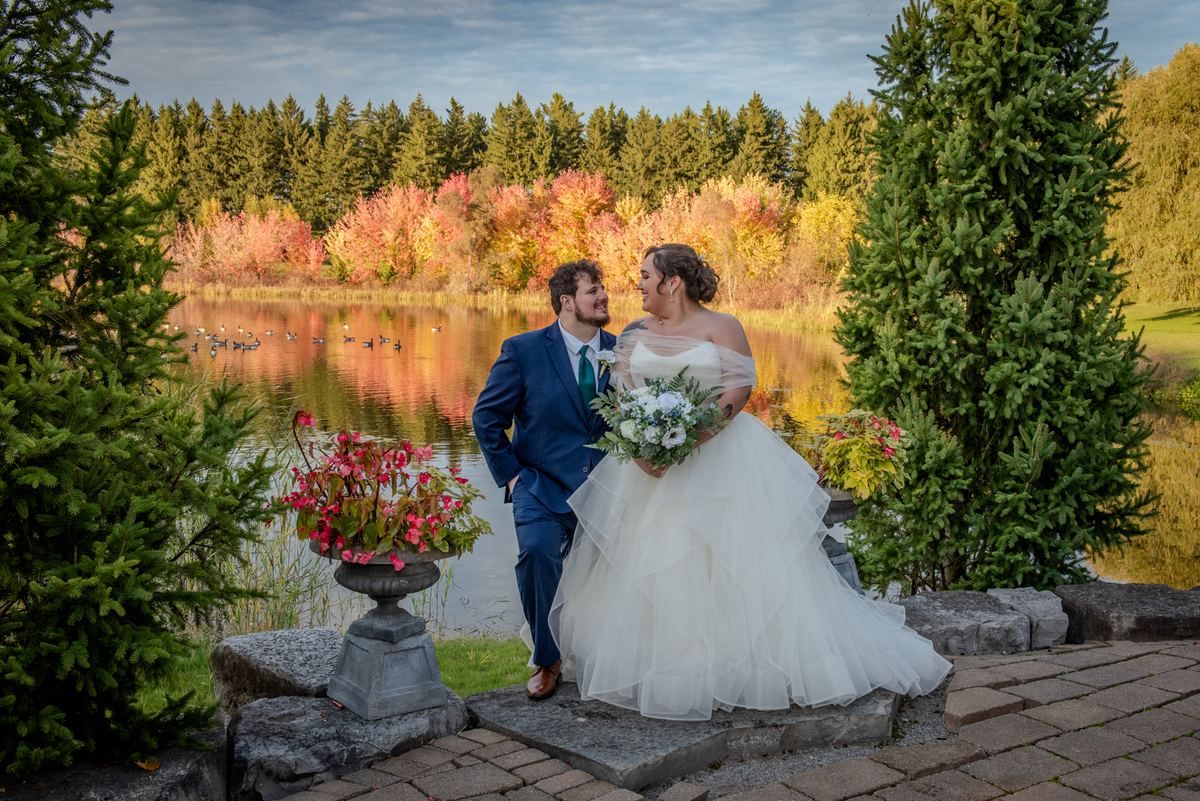A newlywed couple stands on a stone ledge by a serene lake, smiling and holding hands as autumn leaves reflect in the water.