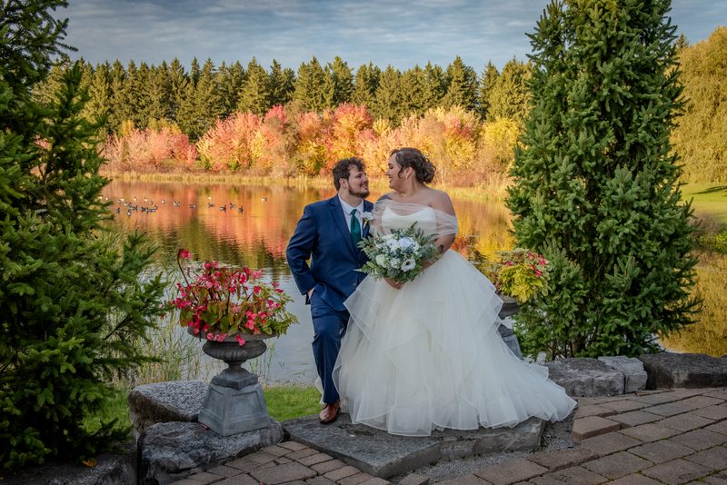 A newlywed couple stands on a stone ledge by a serene lake, smiling and holding hands as autumn leaves reflect in the water.