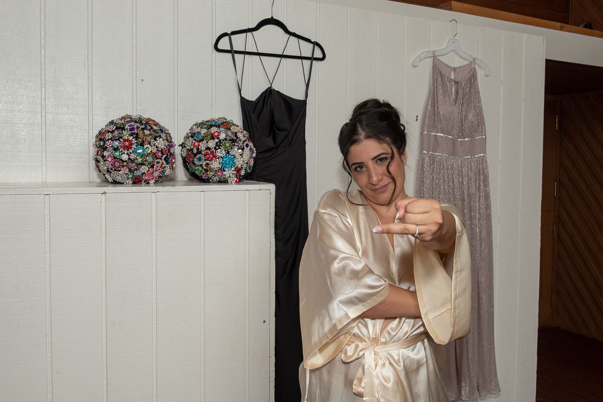 A bride in a satin robe points at her wedding ring, with ornate bouquets and wedding dresses in the background.