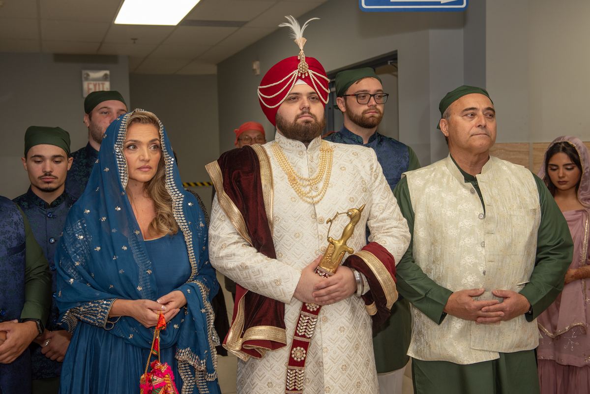 A Sikh groom and his bride in traditional attire stand solemnly with family members in a modern indoor hall, looking upward.
